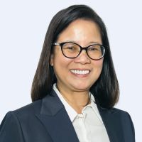 A person with long, dark hair and glasses smiles while wearing a dark blazer and a white shirt against a plain background.