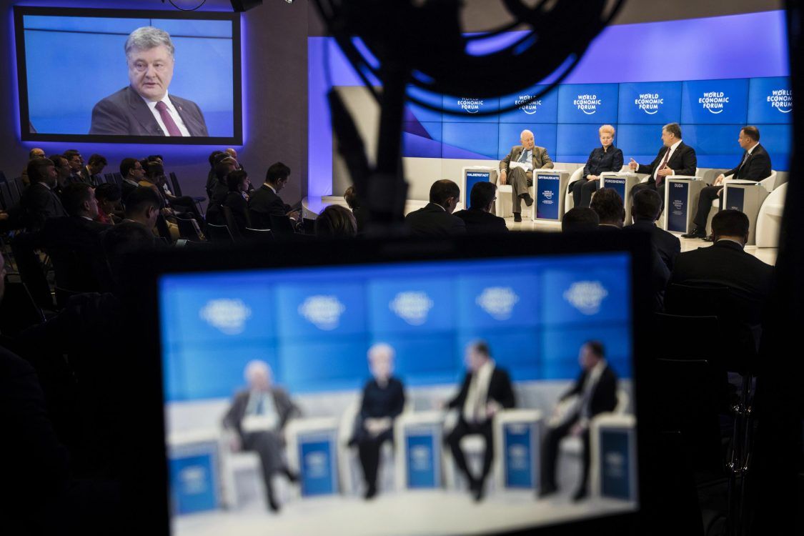 A panel of five people sits on stage at the World Economic Forum, with the discussion shown on a large screen and filmed live for broadcast.