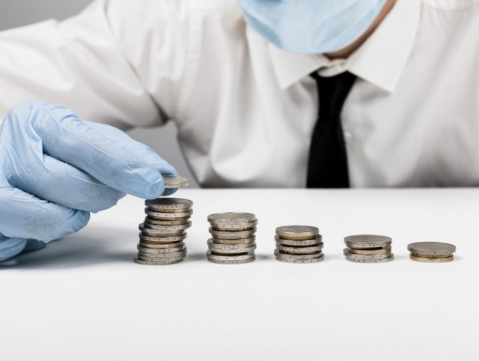 A person wearing gloves and a mask stacks coins on a table, creating columns of varying heights.
