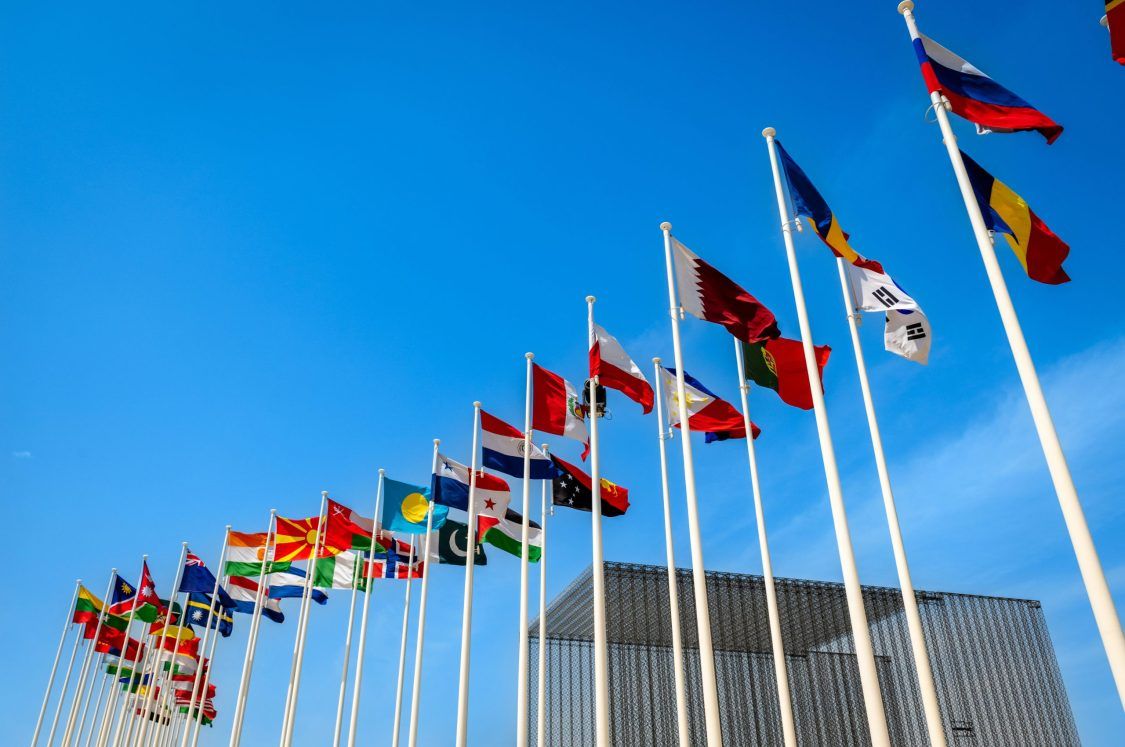A row of national flags from various countries on tall flagpoles against a clear blue sky, with part of a modern building visible in the background.