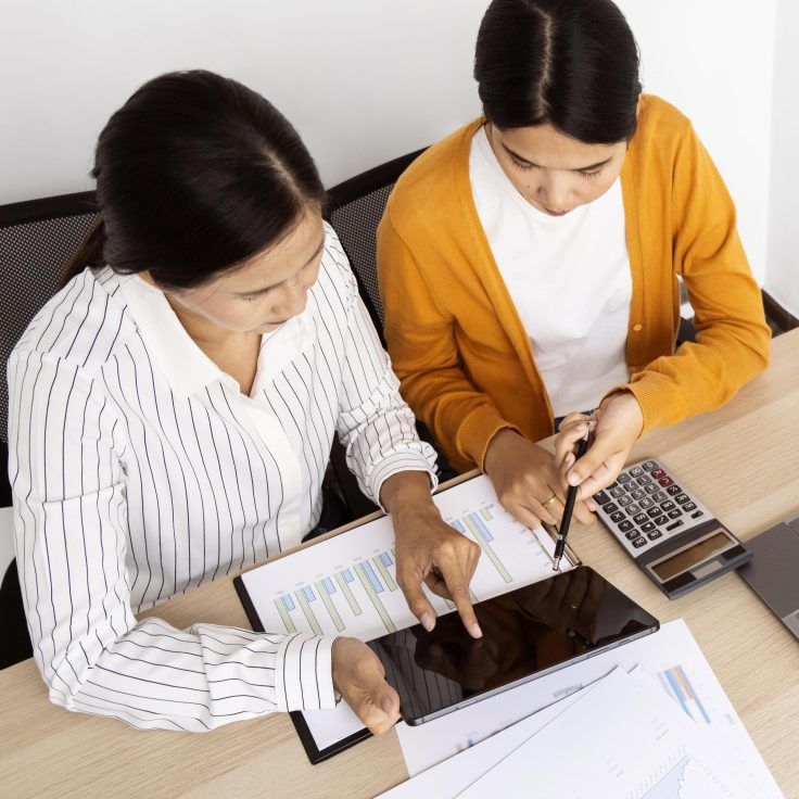Two women working together at a desk, reviewing documents and using a tablet and calculator. One wears a striped shirt while the other wears an orange cardigan. Various charts and graphs are visible.