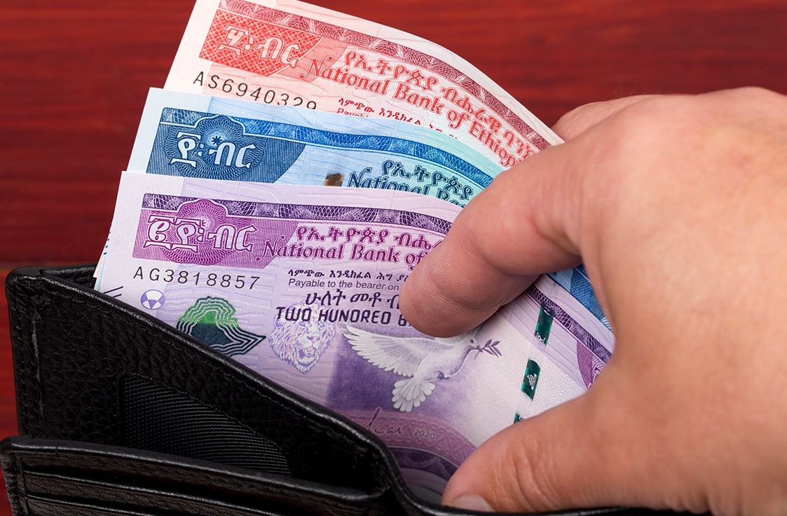 A hand pulls Ethiopian banknotes of various denominations from a black wallet on a red wooden surface.