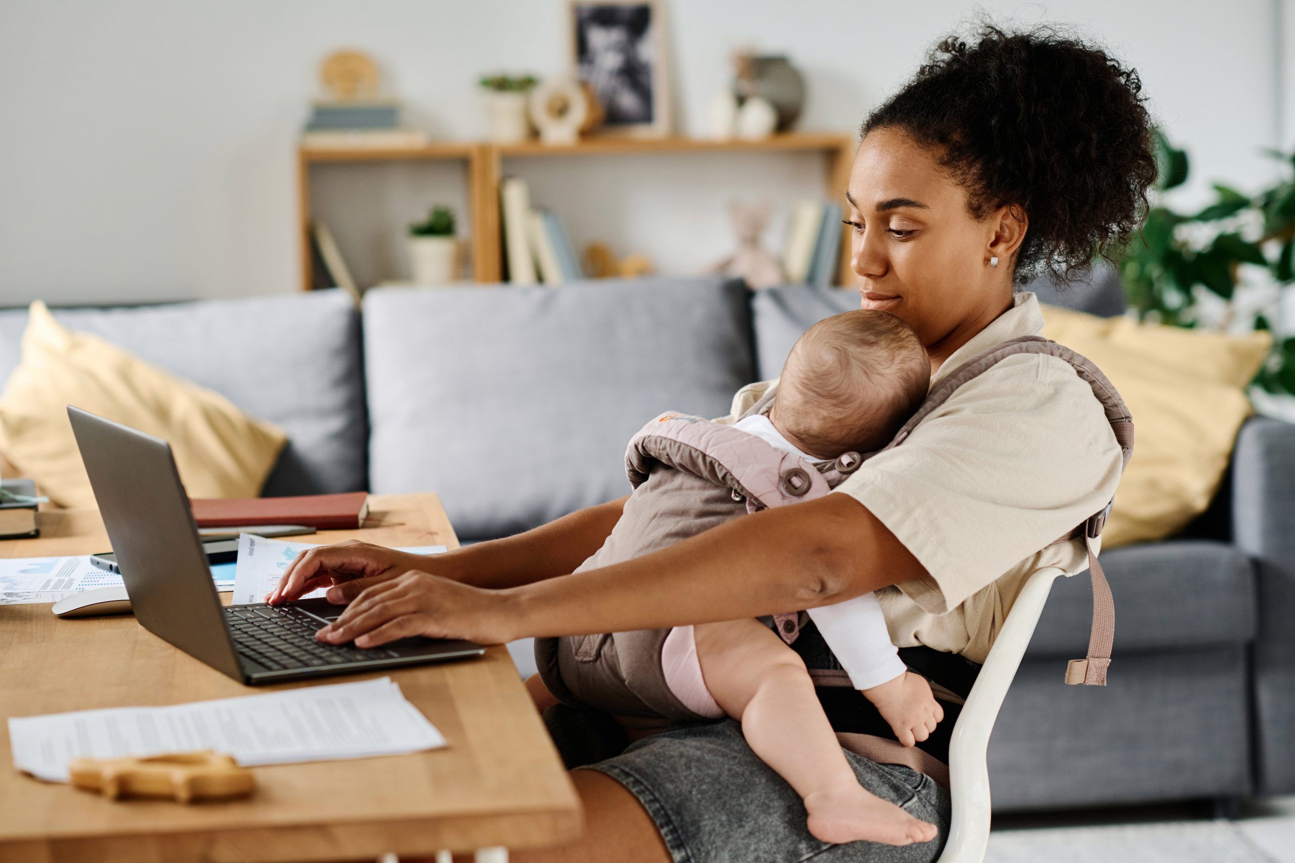 Woman working on a laptop at a desk while holding a baby in a front carrier in a living room setting.
