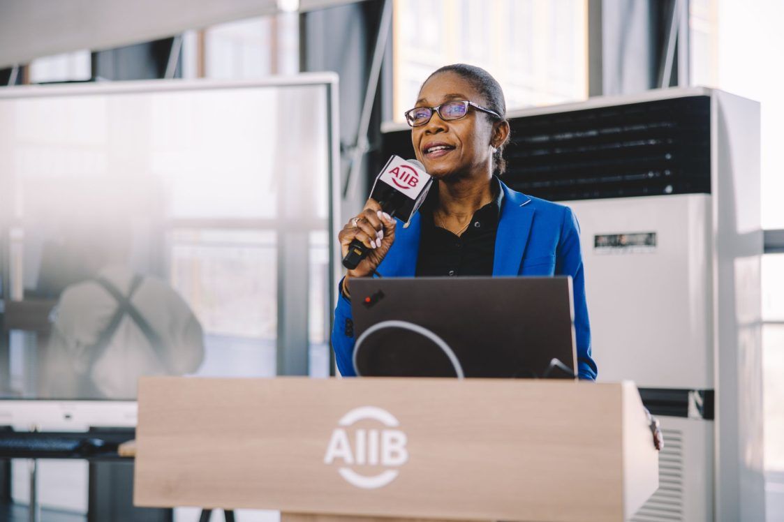 A woman in a blue blazer speaks at a podium labeled AIIB, holding a microphone, with a laptop and whiteboard visible in the background.