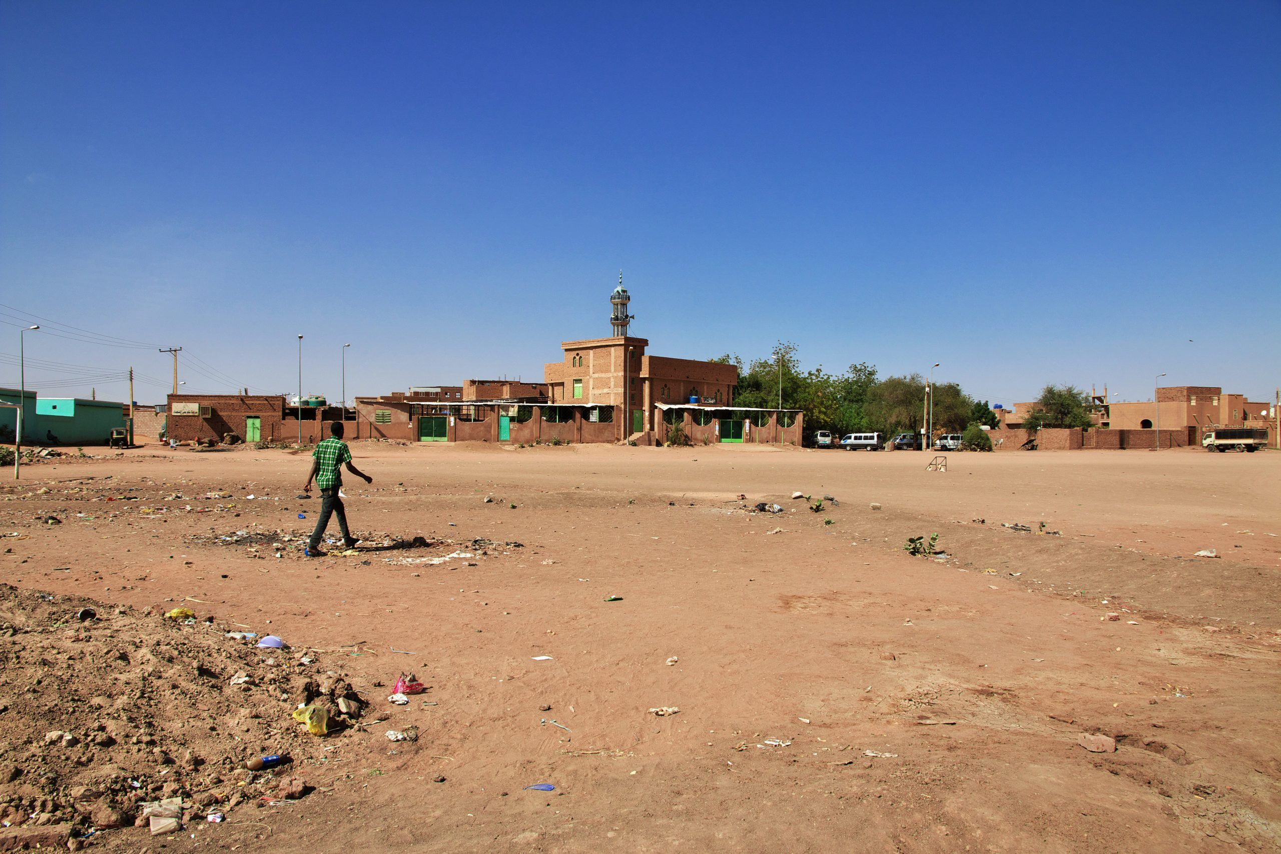 A person walks across a littered, barren dirt field with scattered buildings and a mosque under a clear blue sky in the background.