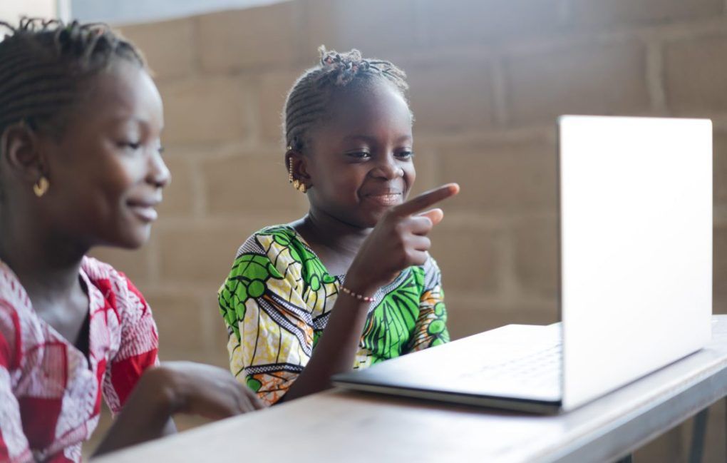 Two young girls sit at a desk, smiling and looking at a laptop screen; one girl is pointing at the screen while the other watches.