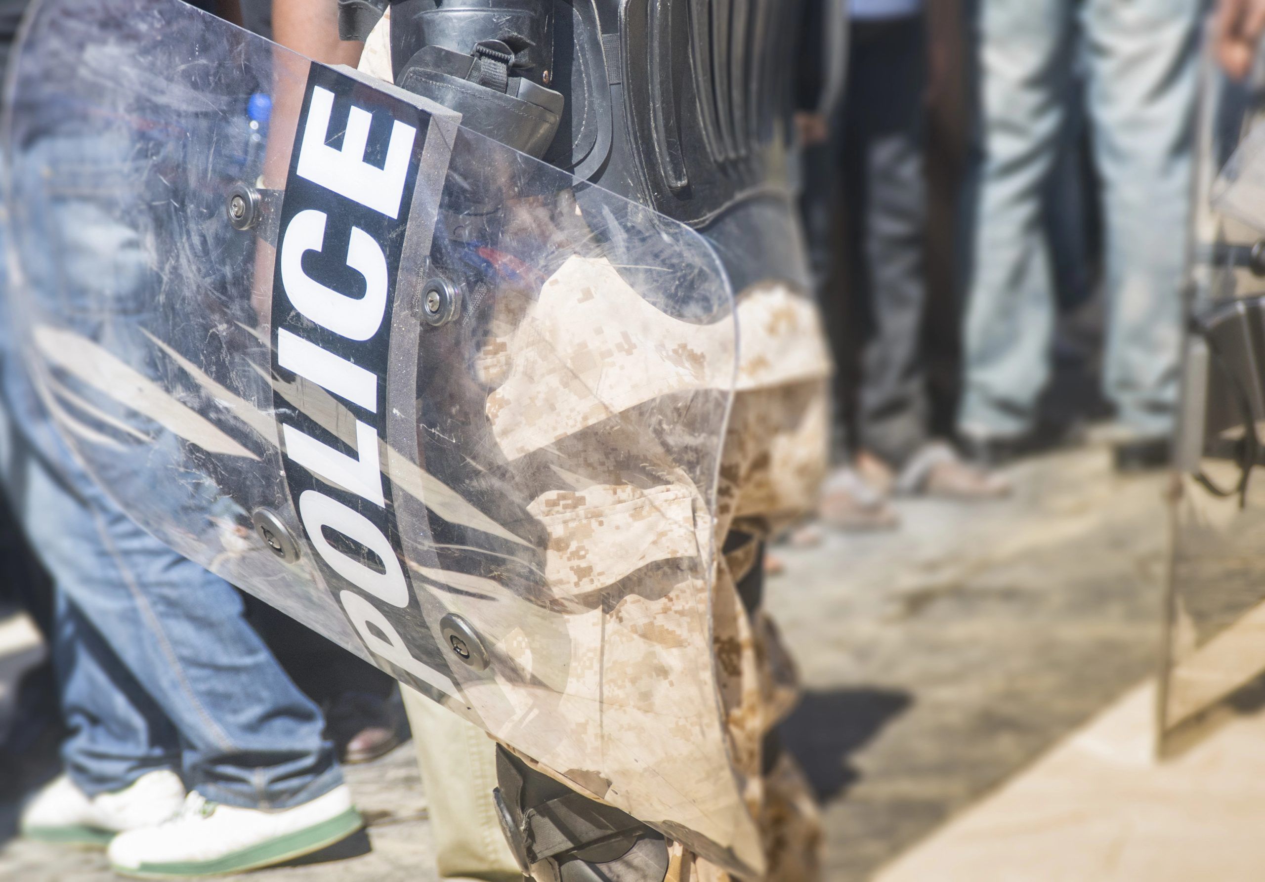 Close-up of a police officer holding a riot shield among a crowd of civilians. The shield has the word "POLICE" written on it. The ground is partially visible, with some debris scattered.