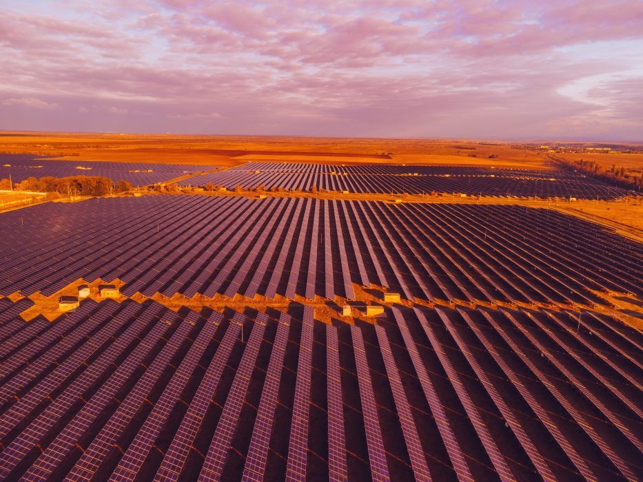Aerial view of a large solar farm with rows of solar panels stretching across a vast, open landscape under a partly cloudy sky at sunset.