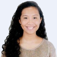 Smiling person with long curly hair wearing a brown sweater against a light background.