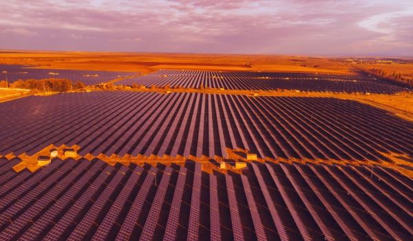 Aerial view of a large solar farm with rows of solar panels stretching across a vast, open landscape under a partly cloudy sky at sunset.