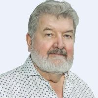 Elderly man with gray hair and beard, wearing a patterned shirt, looking at the camera against a plain background.