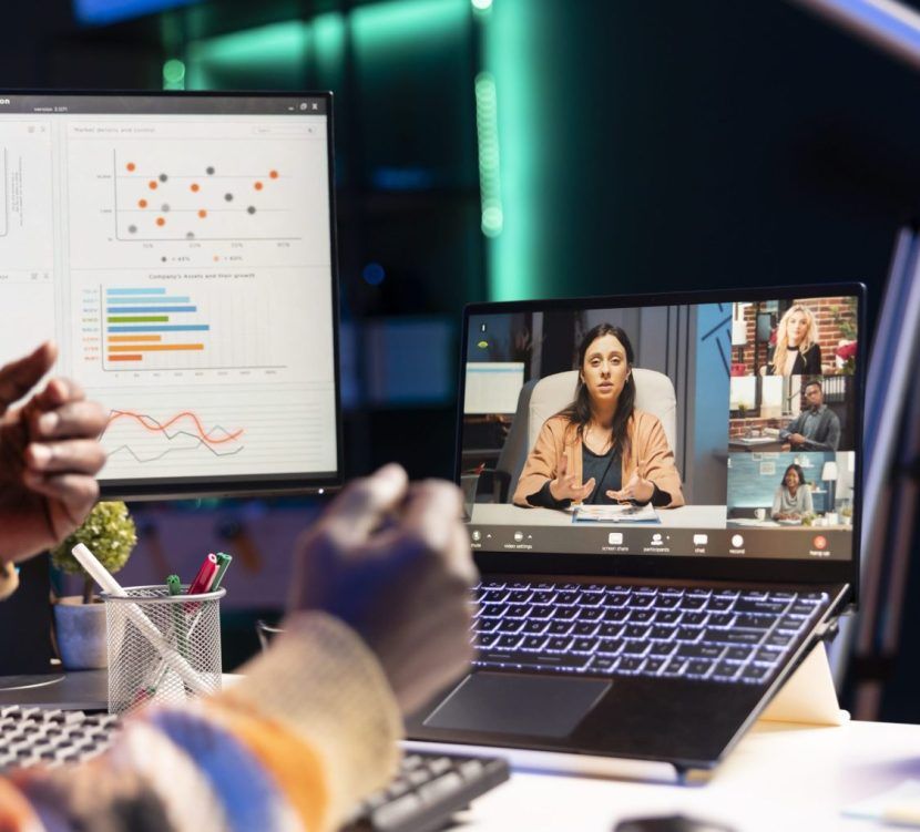 A person participates in a video conference on a laptop, with graphs and charts displayed on a nearby monitor in an office setting.