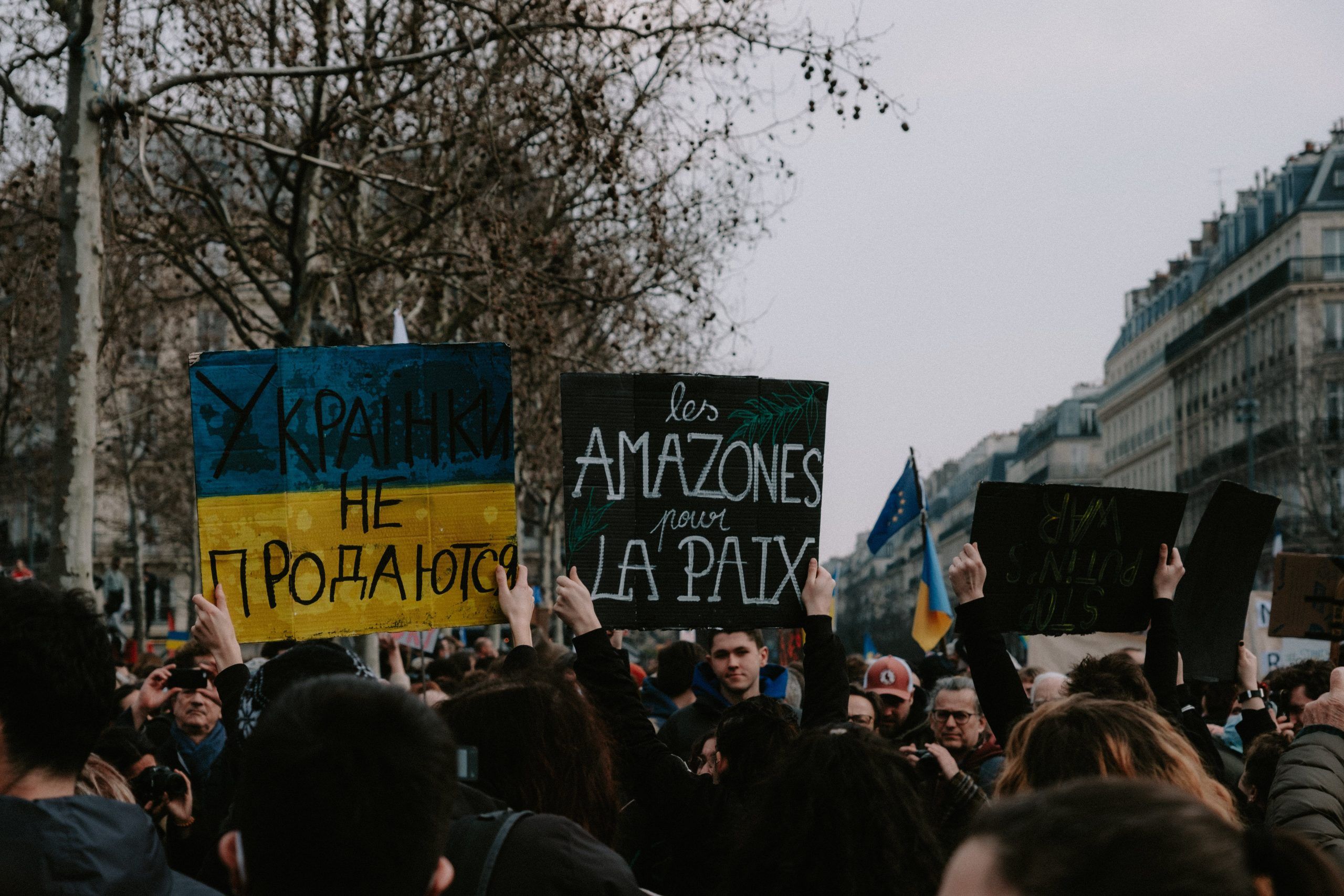 A crowd of people holding protest signs, including a blue and yellow sign in Cyrillic and a French sign calling for peace, gather on a city street with bare trees and buildings in the background.