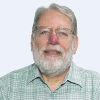An older man with white hair and a beard is smiling. He is wearing glasses and a green plaid shirt.