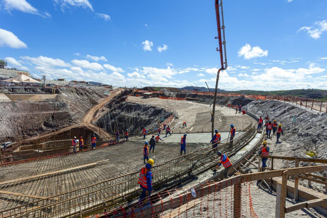 Workers in safety vests and helmets are constructing a large concrete structure at a construction site under a blue sky.