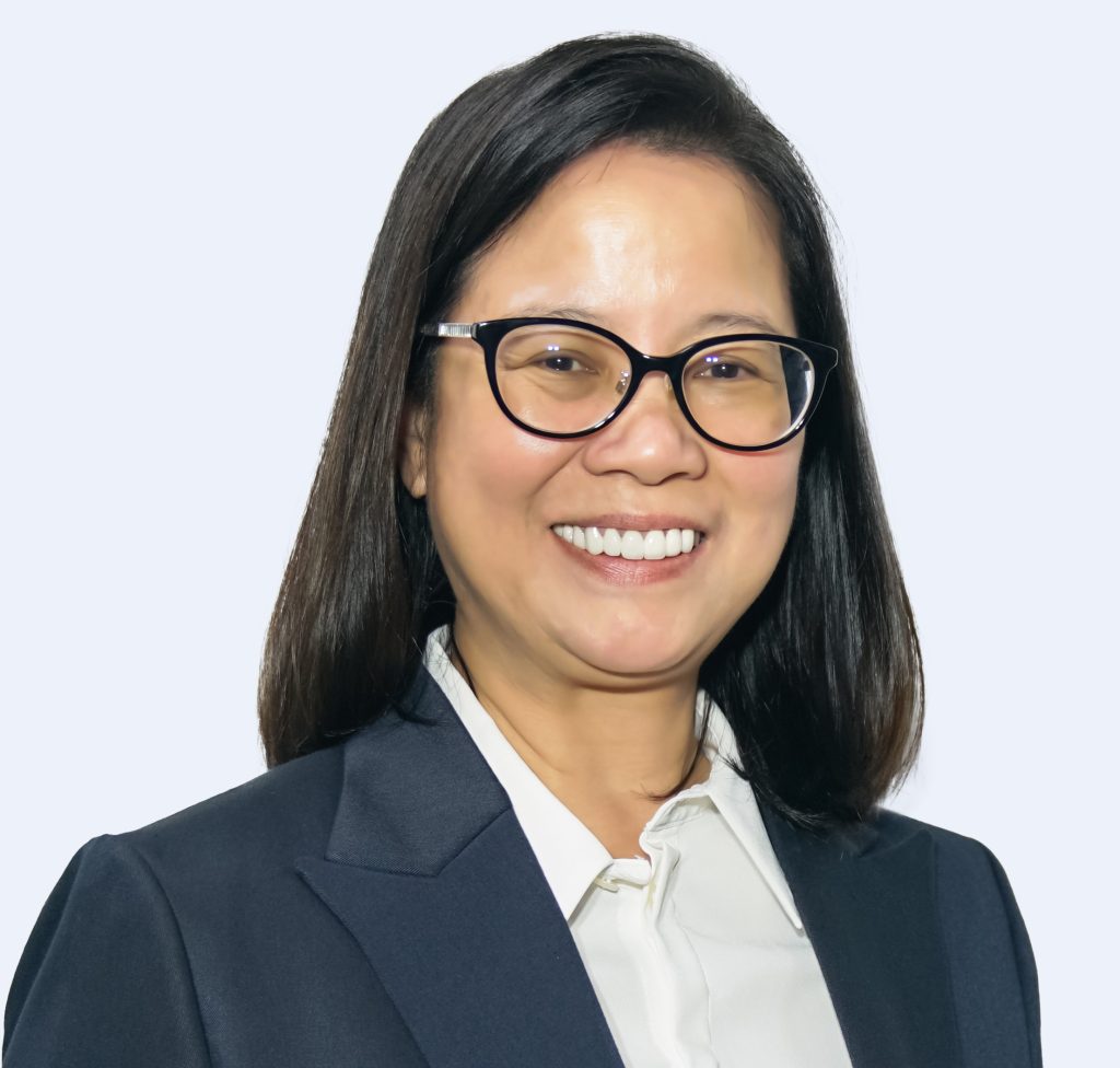 A person with long, dark hair and glasses smiles while wearing a dark blazer and a white shirt against a plain background.