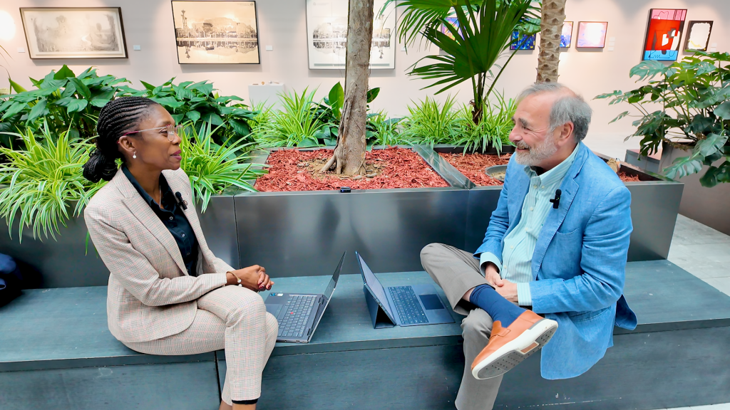 Two people sit on a bench indoors with laptops, engaged in conversation, surrounded by plants and framed photos on the wall in the background.