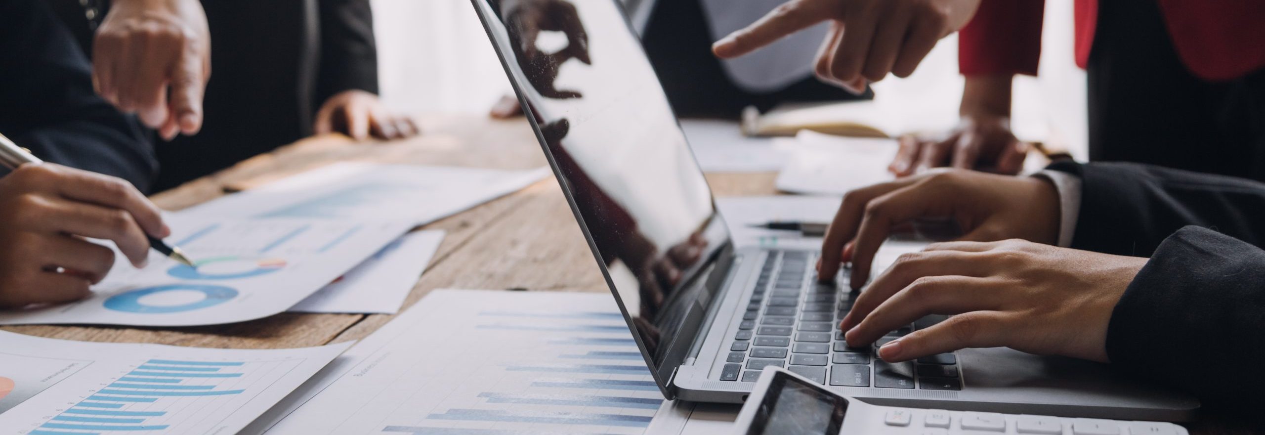 People working at a desk with a laptop, calculator, and papers displaying graphs and charts. Several hands are pointing and typing, suggesting a collaborative work session.