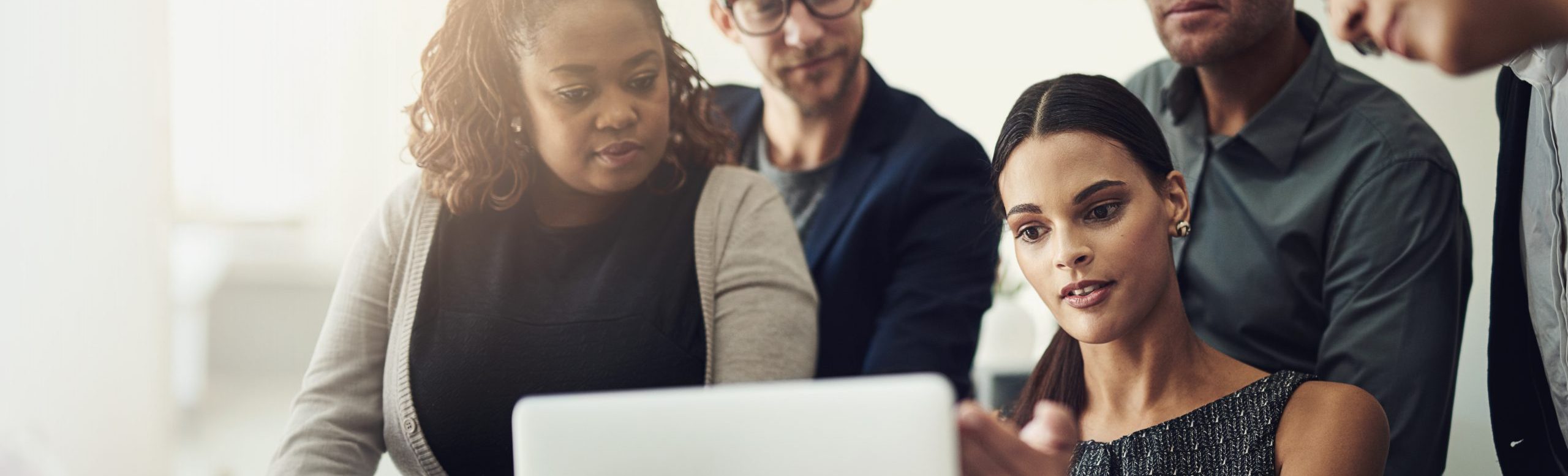 Five people gather closely around a laptop, attentively looking at the screen while one woman gestures as she explains something to the group.