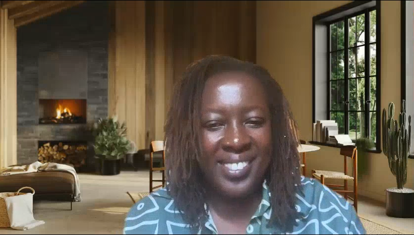 A woman smiling at the camera is seated in a modern, warmly lit room with wooden walls, a fireplace, large window, and plants.
