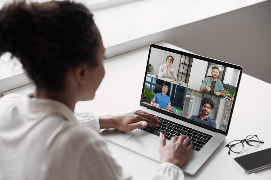 A person sits at a desk participating in a video conference on a laptop with four other people. Glasses and a smartphone are on the desk.