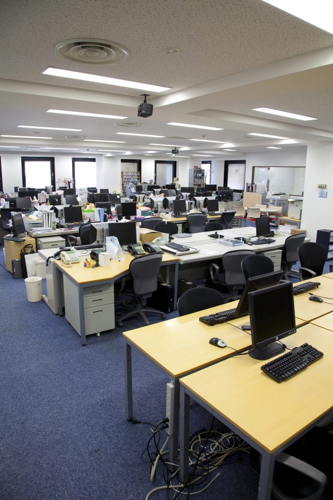 A spacious, modern office with rows of empty desks, computers, office chairs, and various office supplies, under bright fluorescent lighting.