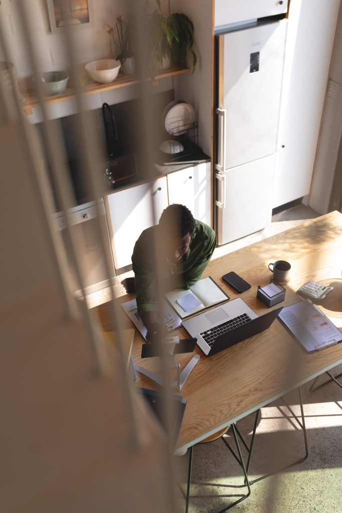 A person sits at a kitchen table working on a laptop with papers, notebooks, a mug, and a smartphone scattered around. The scene is lit by natural daylight.