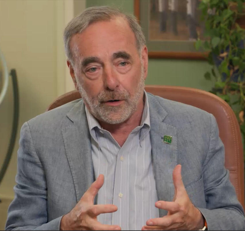 An older man with gray hair and a beard, wearing a light blue blazer and a striped shirt, sits in an office chair and gestures with his hands while speaking.